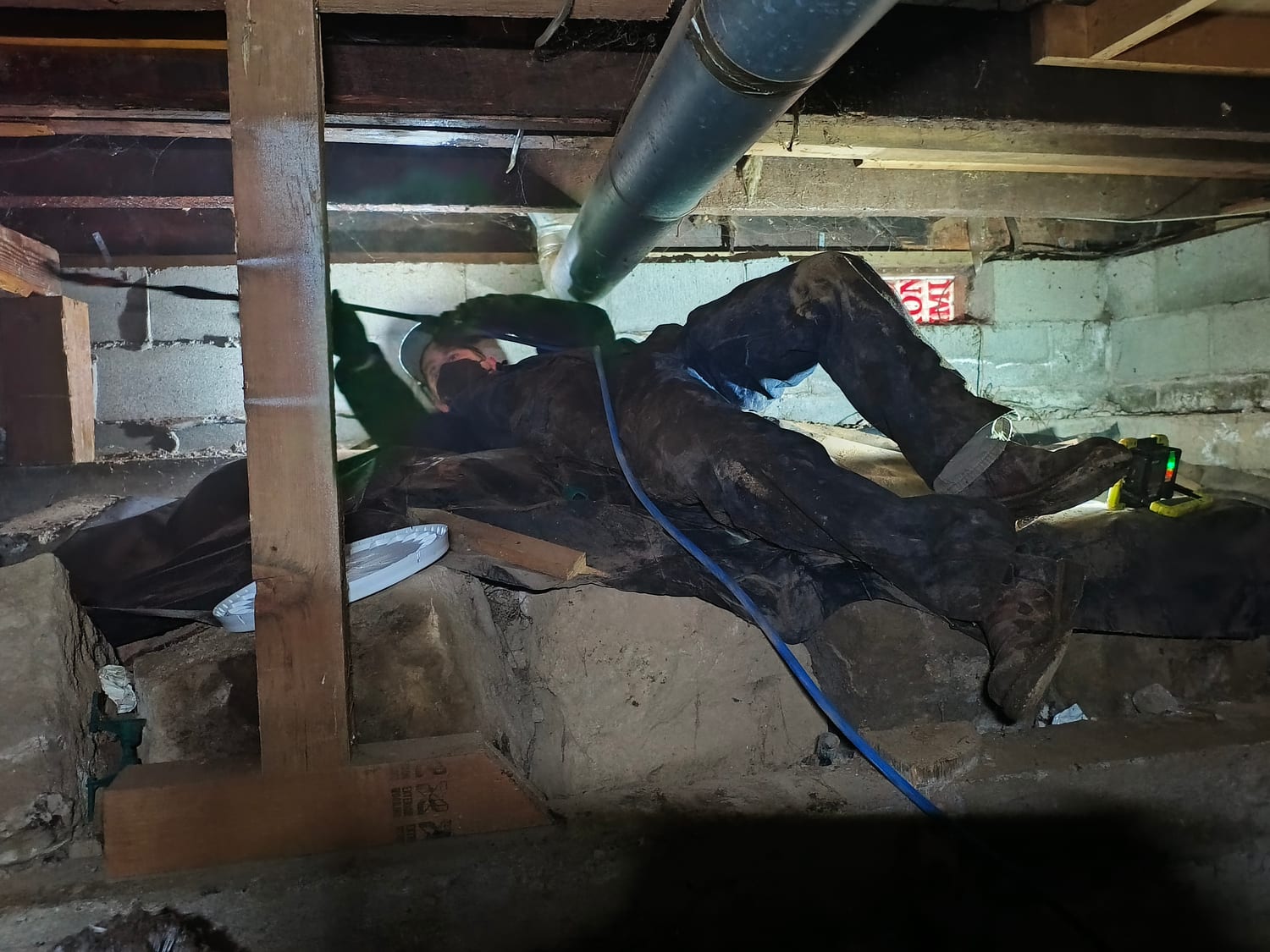 A technician in coveralls, a respirator and a headlamp lying on his back on a crawlspace vapor barrier, spraying the joists overhead, with a blue hose running to the sprayer
