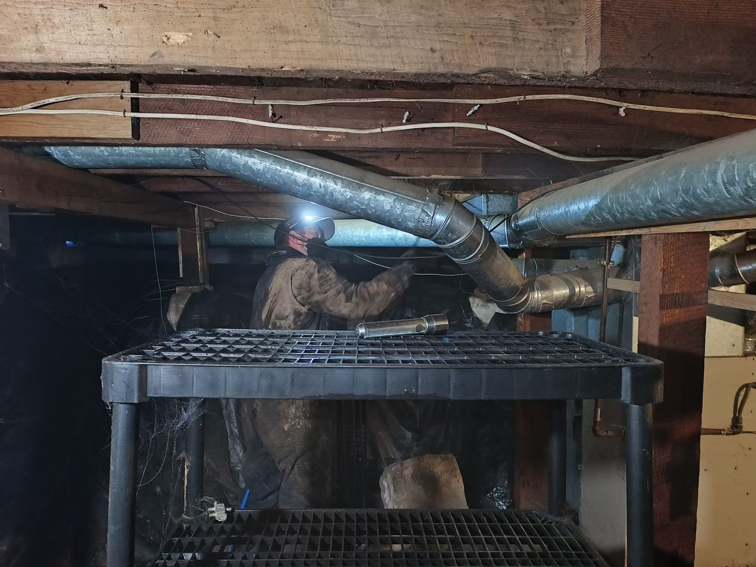 Travis (TJ) Hansen by headlamp in a crawlspace in Burlington, working under the ducting with a sprayer wand